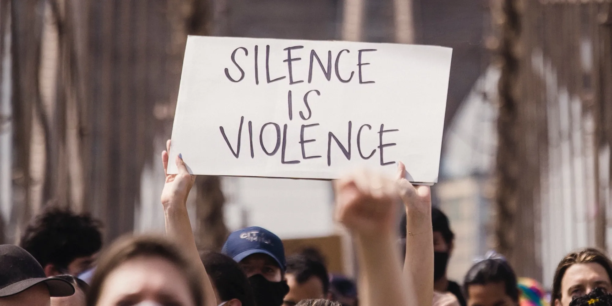 A crowd is walking across the Brooklyn Bridge. One protestor holds up a sign that reads, "Silence is Violence."