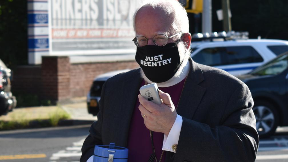 The Rt. Rev. Andrew M.L. Dietsche stands in front of the Rikers Island sign with a megaphone and a mask reading "Just Reentry."