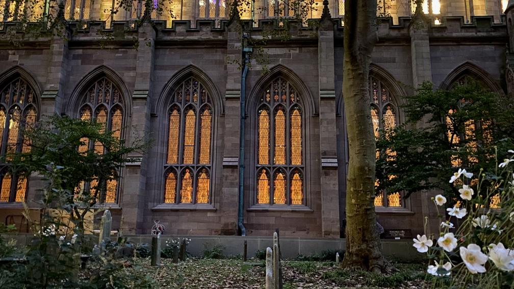 Golden light shines from within Trinity Church and lights up the windows, as seen from Trinity Churchyard on an autumn evening