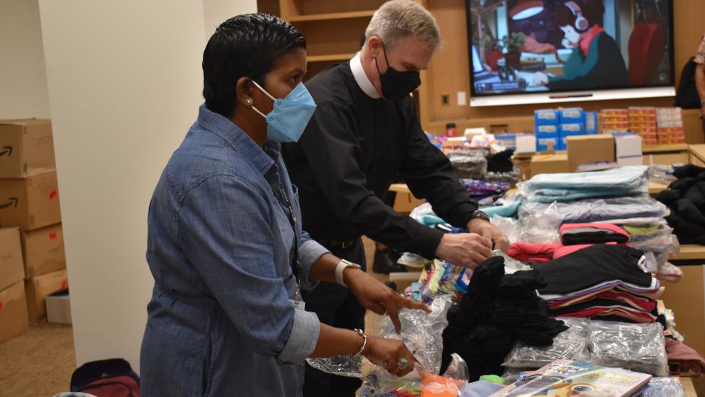 A brown skinned woman with short hair wearing a blue mask & white man wearing priest's garb and a mask pack books, clothing, and other supplies. The room is full of boxes and boxed food items.