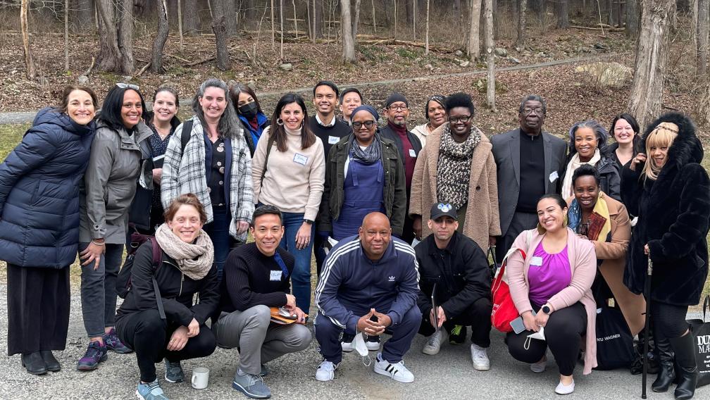 A multiethnic group of people wearing coats, scarves, and other outerwear smile for a group photo outside the Trinity Woods in West Cornwall, Connecticut.