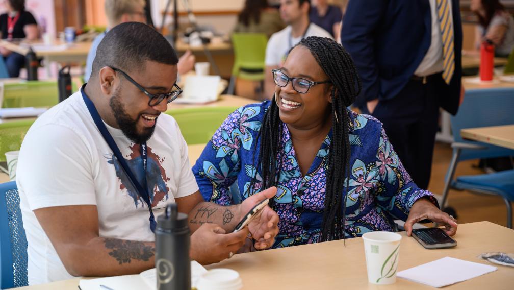 Two people are laughing together during paired conversation. The Black man wears a white tee with a graphic print, and glasses, while the Black woman has braided hair and wears a blue floral dress and glasses.