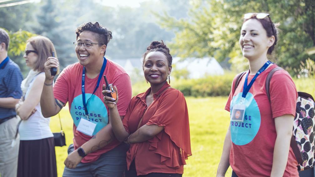 Members of Princeton Theological Seminary's Log College Project wear red shirts and smile at an unseen group. In the foreground is a young  person of color with tattoos and glasses, standing next to a smiling Black woman looking directly at the camera. A young white woman wearing a white and black spotted backpack in another red shirt with blue dot logo that says "Log College Project" also smiles at the camera, while slightly blurred in the shot.