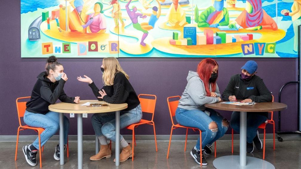 Masked young people sit in pairs at round tables having conversations. A colorful mural that says "The Door" along with words like "support," "acceptance," "love," "welcome," "unity," "ally," and community."