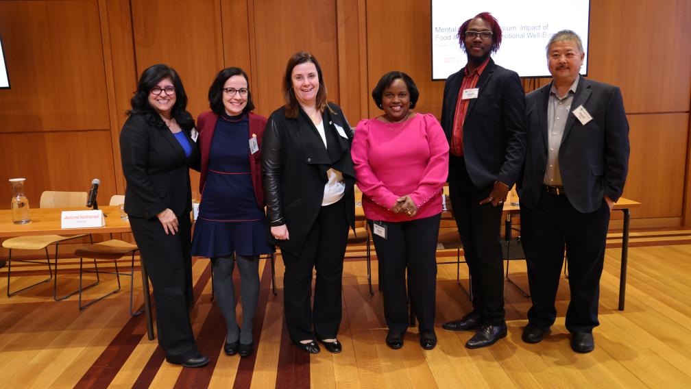 Six panelists of varying ethnic and racial background stand in a row, smiling at the camera. The screen behind them says "Mental Health Symposium: Impact of Food Insecurity on Emotional Well-Being"