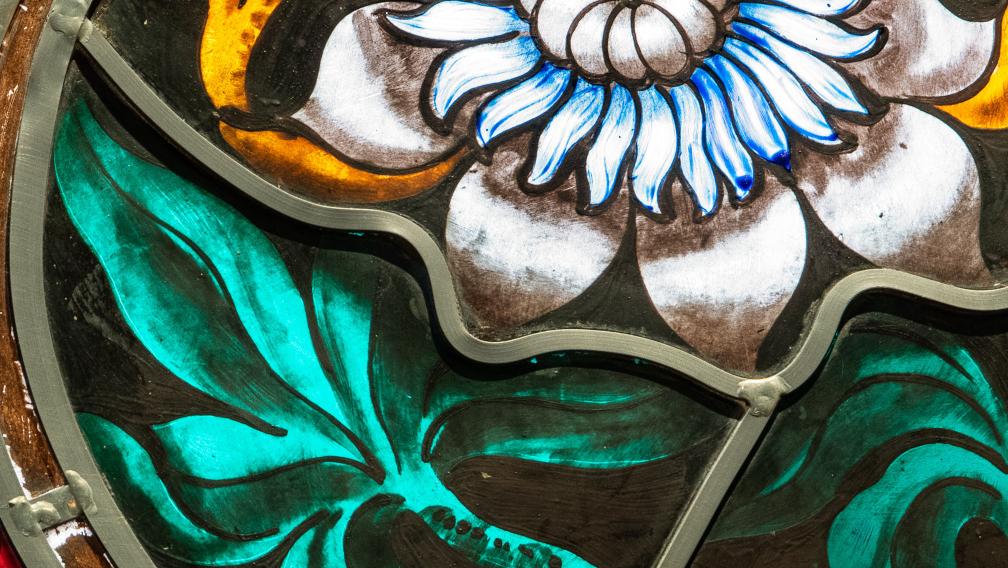 A close-up photo of stained glass depicting a white and blue flower with vivid green leaves