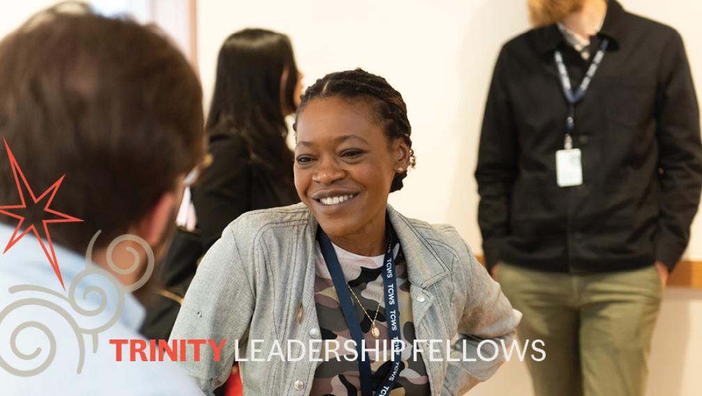 A group of Trinity Leadership Fellows stand in conversation. Chiseche Mibenge smiles at her colleague whose back is facing us. The text "Trinity Leadership Fellows" is overlaid over the photo, with a red six point star and grey swirling clouds at left.