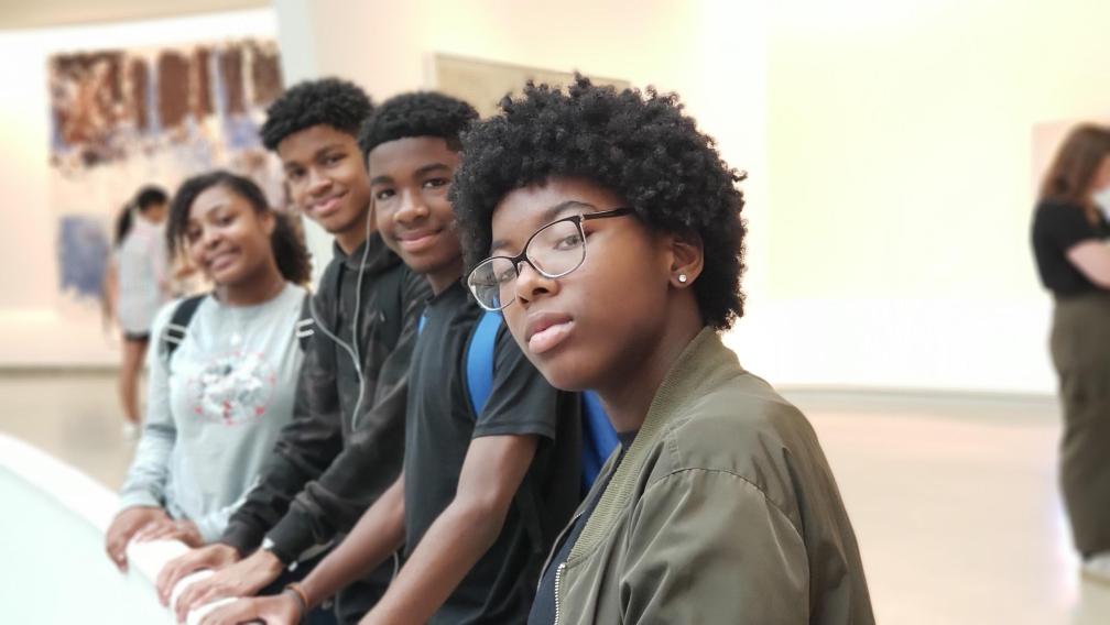 Four black children stand in a row facing the camera. The three children in the background smile softly, while the child in the foreground wears glasses and looks seriously at the camera.