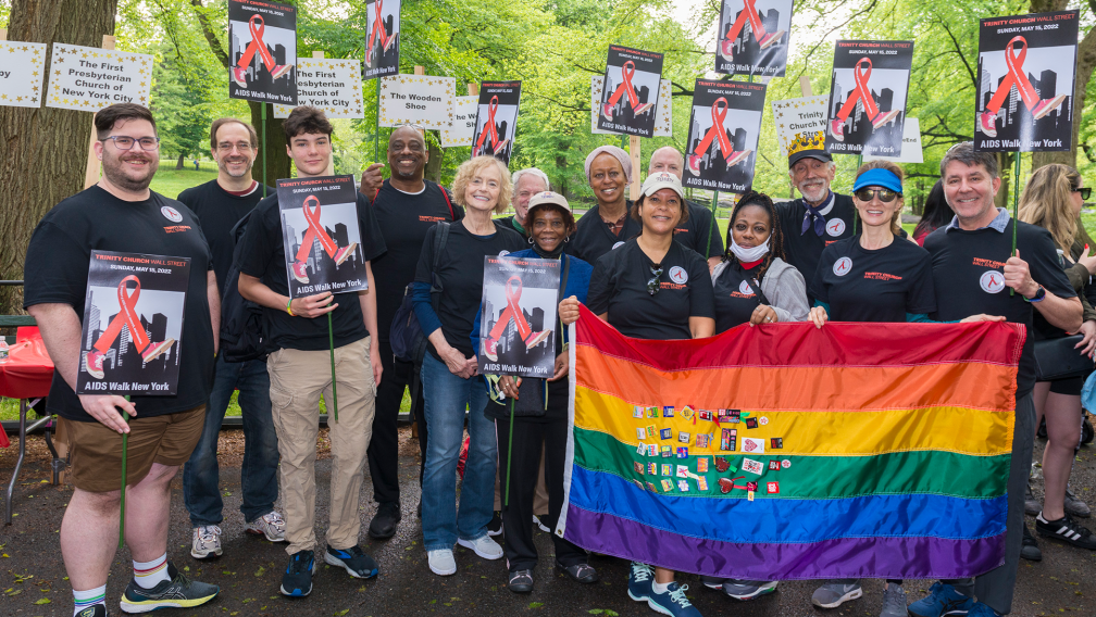 A group of people from Trinity Church Wall Street stand holding signs with the AIDS red ribbon and a Pride flag, at the AIDS Walk 2022