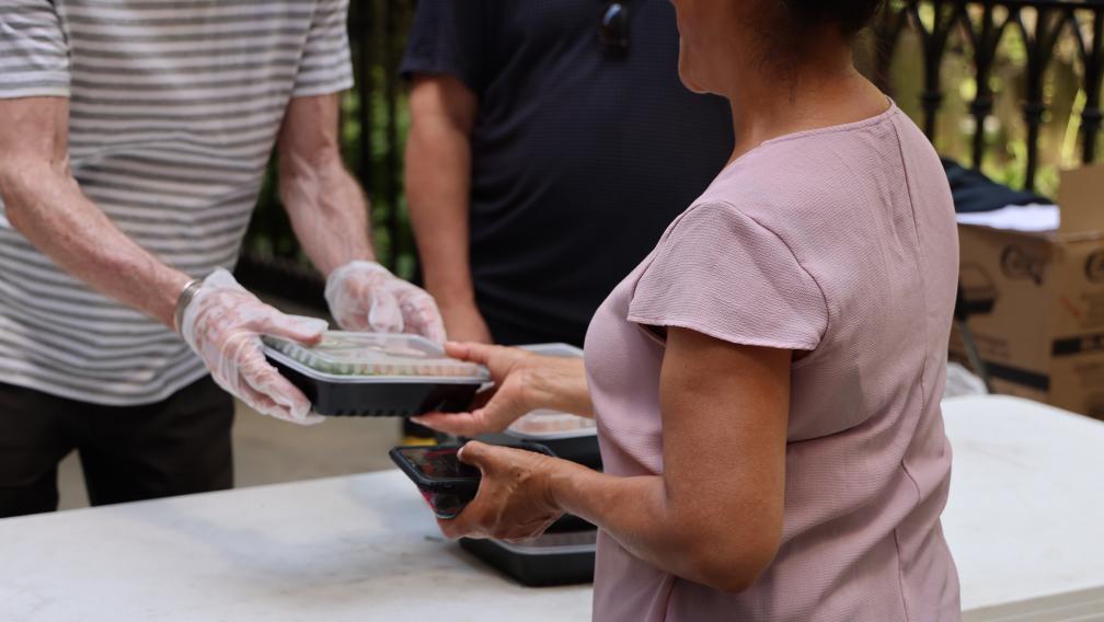 A gloved volunteer hands a Compassion Meal to a guest in Trinity Churchyard.