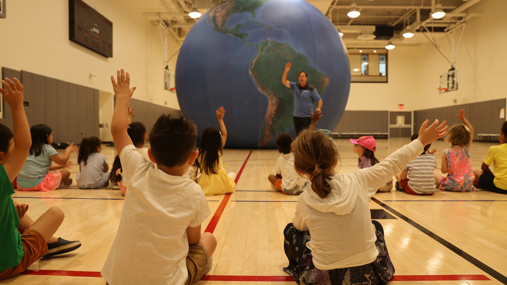 Children in the Trinity Commons Gym raise their hands in front of an inflatable globe as part of a science class during the 2023 Children's Summer Program.