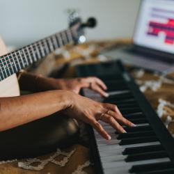 A person holds a guitar and plays a keyboard with a laptop in the background