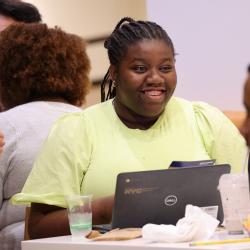 Student smiles across table. She wears a lemon colored short sleeved top, and her braided hair is pulled back into a pony tail. 