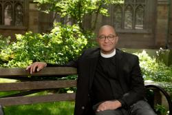 The Rev. Phillip A. Jackson, Rector sits on a wooden bench in the Trinity Churchyard. He has closely cropped hair, round, horn-rimmed glasses and is wearing a clerical collar under a black jacket.
