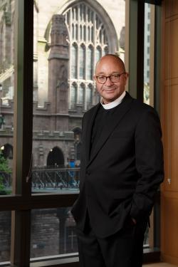 The Rev. Phillip A. Jackson, Rector stands in his office, which overlooks Trinity Church. He has closely cropped hair, round, horn-rimmed glasses and is wearing a clerical collar under a black jacket.