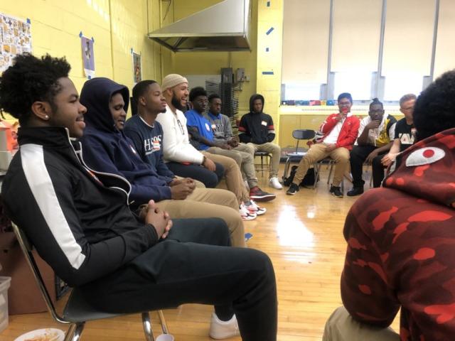 Young Black men sit in a circle facing each other in a classroom. They are smiling.