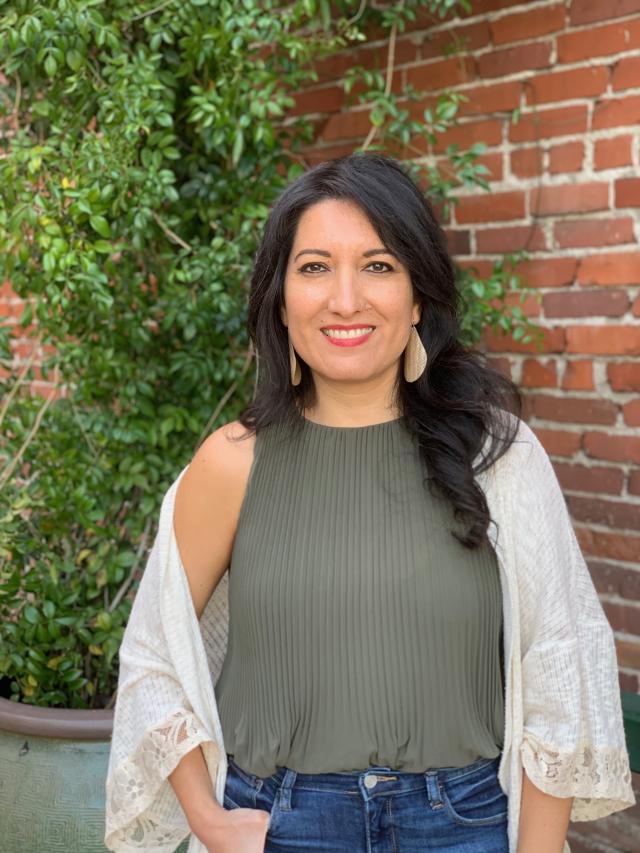 Inés Velasquez-McBryde smiles into the camera, standing in front of a brick wall and shrubbery. She wears a green tank, blue jeans, white shawl, and dangly earrings.