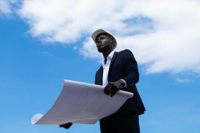 A Black man wearing a hard hat looks up towards the sky. He holds blue prints in his hand, as the camera is angled from below.