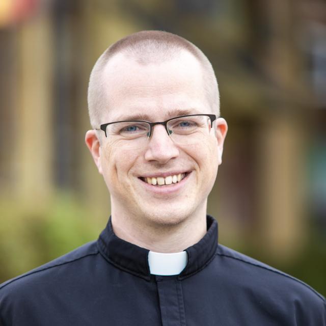Rev. Ben Groth smiles directly at the camera, wearing a priest's collar and glasses.