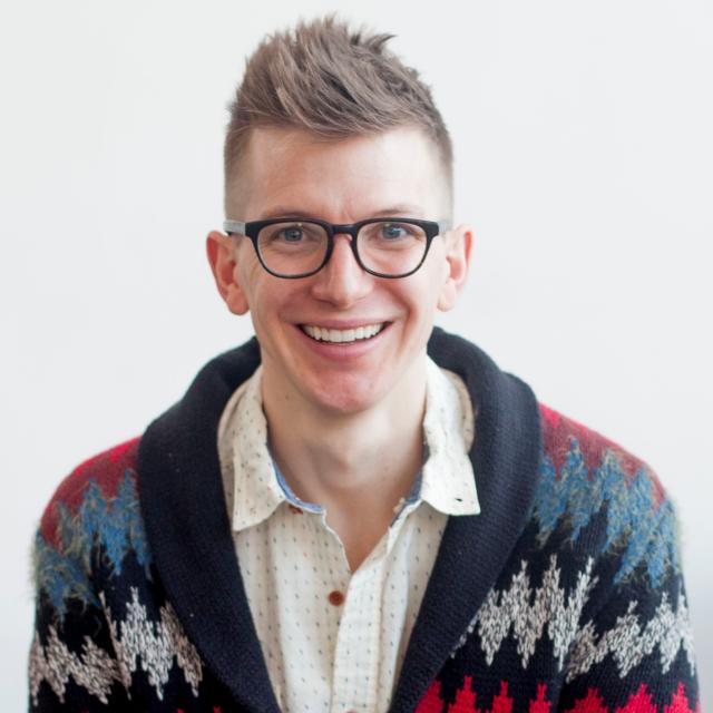 David Washer faces the camera, smiling widely, while wearing glasses, a patterned button down and cardigan.