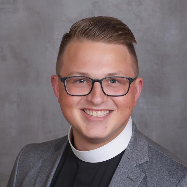 The Rev. Isaac Petty smiles directly at the camera, wearing a priest's collar, grey blazer, and glasses. He stands in front of a grey background.