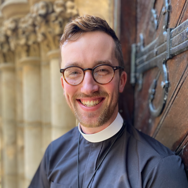 The Rev. Kevin Gordan Neill smiles broadly at the camera, wearing clerical attire and glasses. He stands in front of wooden church doors and stone columns.