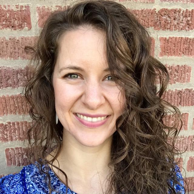 The Rev. Lauren Grubaugh Thomas smiles directly at the camera, wearing a blue paisley blouse as she stands in front of a brick wall.