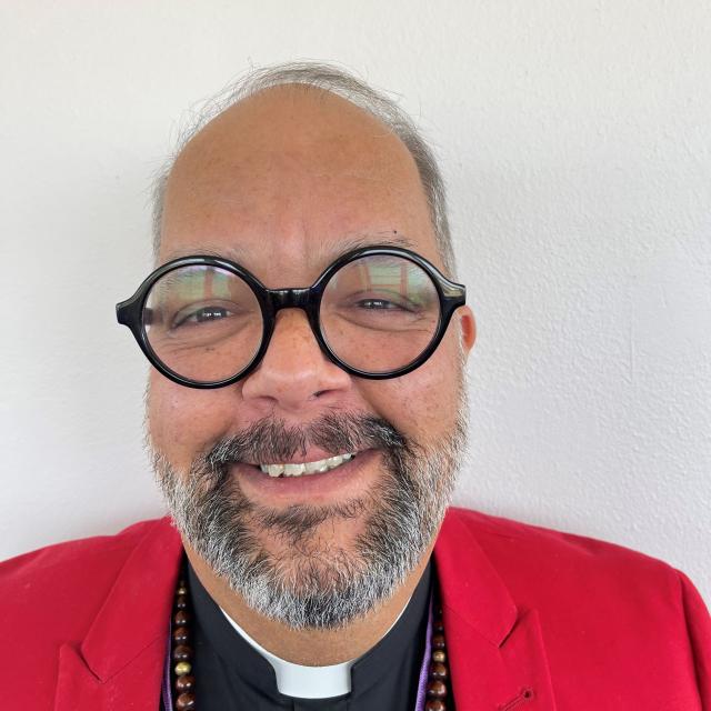 The Rev. Peter Vázquez-Schmitt smiles into the camera, wearing a red jacket, round glasses, and a priest's collar.