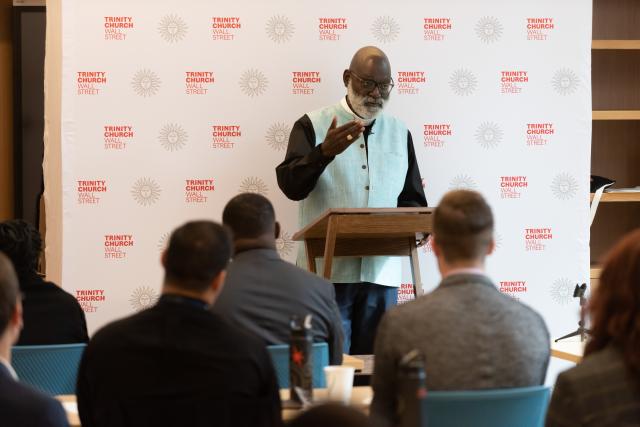 Father Mark wearing a light blue vest and priest collar, address a group of Trinity Leadership Fellows. He stands in front of a step and repeat featuring Trinity Church Wall Street branding.