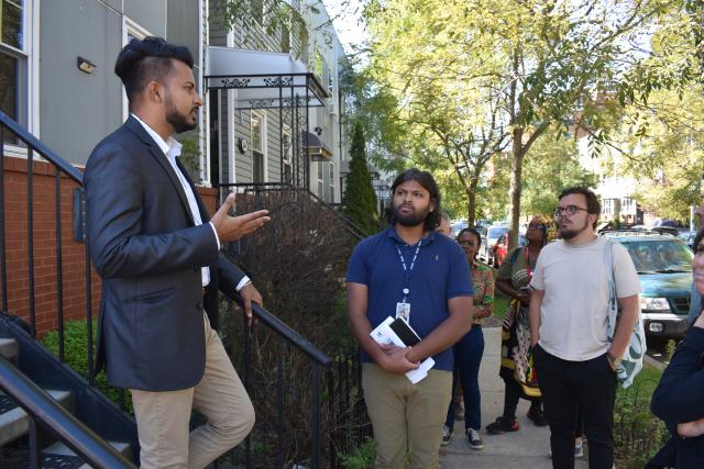 A South Asian man in a navy blazer & khaki pants stands on a staircase while addressing Trinity Leadership Fellows outside. The Fellows are dressed casually while standing in residential Brooklyn.