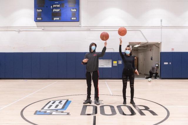 Two masked young people dressed in all black spin basketballs on their fingers while standing on a basketball court. The decal on the court reads "The Door."