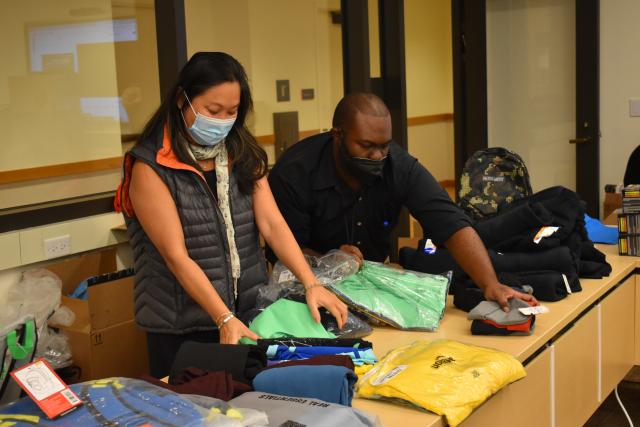 An Asian woman and Black man, both masked, pack clothes & other supplies for asylum seekers. They are surrounded by clothes, bags, and boxes..