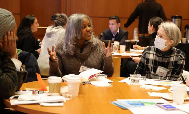 In the foreground, women of different ages sit at table in conversation during the Mental Health Symposium. The woman on the right wears a mask and a plaid top; the woman in the center wears a grey shawl with her grey hair down in waves. The woman at left wears a headwrap with her face cradled in her right hand.