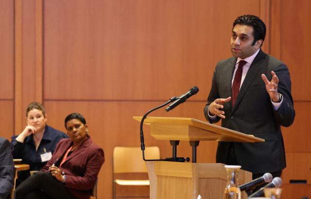 Dr. Ashwin Vasan wears a dark suit with light pink collared shirt and red tie, addressing an audience. Lorelei Vargas and Vidia Cordero sit in the background, wearing professional attire.