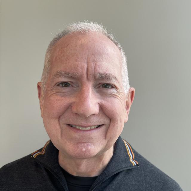 Thomas Armstrong, an older white man, smiles into the camera. He has white hair and is clean-shaven, and is wearing a black tee under a grey half-zip sweater with striped accents on the collar.