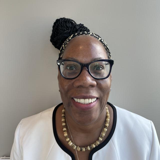 Cecilia Scott-Croff smiles, wearing black glasses, a black patterned headband, and her hair in a bun. She wears a neutral beaded neckace and white collarless jacket with black trim.