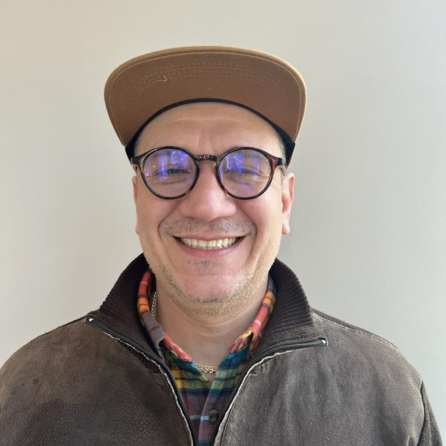 Frank Gonzalez smiles at the camera, wearing a tan cap, round glasses, plaid shirt, and brown jacket.