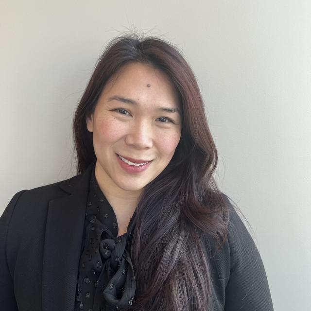 Eva Wong smiles at the camera, freckled cheeks rosy and pink lipstick. She wears a tonal black polka dot blouse with a bow at the collar under a black blazer. Eva is of East Asian descent with burgundy tinted hair, swept to the side and worn down.