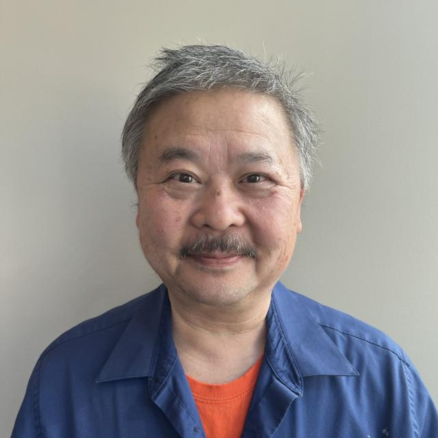 Don Hong smiles at the camera, wearing an orange tee under a blue collared shirt. He is of East Asian descent, with greying hair and a mustache.