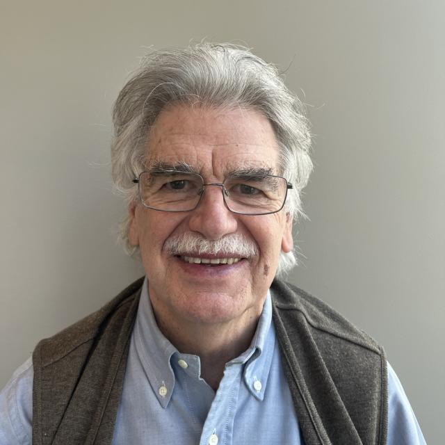 Jaime Inclán smiles at the camera, wearing a wire-framed glasses and grey vest over a blue button-down shirt. He has white hair and mustache.