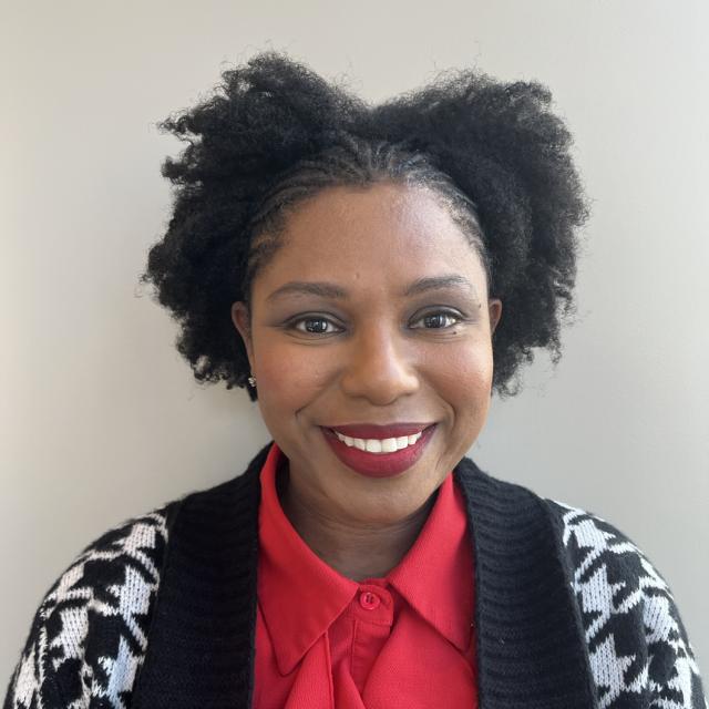 Tiffany Winbush smiles at the camera, wearing red lipstick, a matching red blouse, and black and white houndstooth cardigan. She is a Black woman wearing her hair braided in the front with her natural hair out mid-crown.