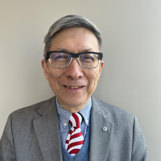 Wellington Chen smiles widely, wearing two-toned glasses, a red and white striped tie, blue checkered button-down, blue vest, and grey checked blazer. He is an elderly East Asian man with freckles and grey short-cropped hair.