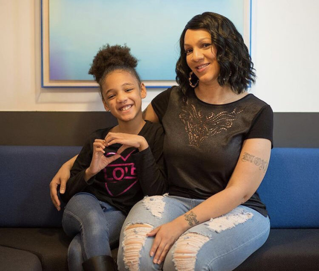 A young Latina girl and her mother smile at the camera, both wearing black tops and denim pants. The daughter's hair is in a high ponytail poof and she makes the heart symbol with her hands. The mother, who has wavy dark hair and a few arm tattoos, wraps her arm around her daughter.