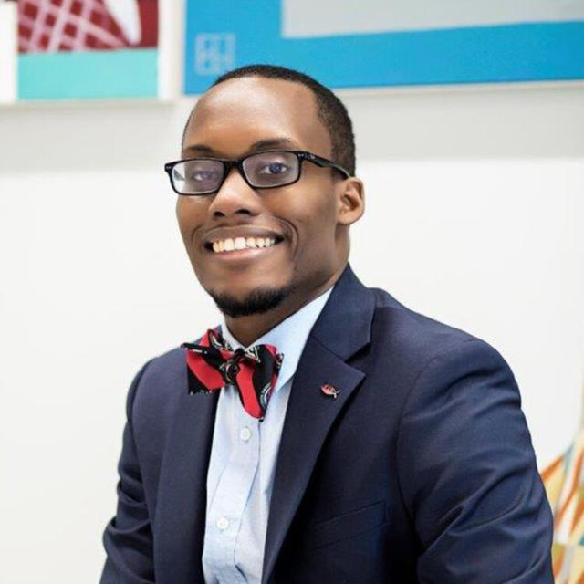 Aaron Rogers smiles broadly at the camera as he sits in front of a white backdrop with colorful paintings out of focus. He is a black man with dark hair, dark eyes, a goatee, and deep brown complexion. He wears a red and navy striped bowtie, light blue button up shirt, and navy blazer with a pin at his lapel.