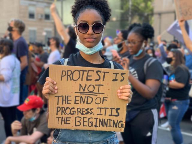 A BroSis youth organizer holds a sign that reads "Protest is not the end of progress. It is the beginning." She wears dark round sunglasses and a blue mask pulled down under her chin. Her dark curly hair is pulled into a puff at the crown of her head.