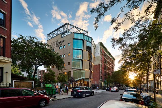 An external shot of The Brotherhood Sister Sol's new building on West 143rd street. The sun sets in the background, shining through trees that dot a car-lined street full of residential buildings. The sky is blue with some white clouds dotting the skyline.