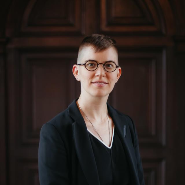 Flourish Klinke, a non-binary White person with short hair, pale skin with a golden undertone, stands in front of a dark wood door. They wear a black blazer and black dress with dark round glasses and silver necklace tucked into their neckline.