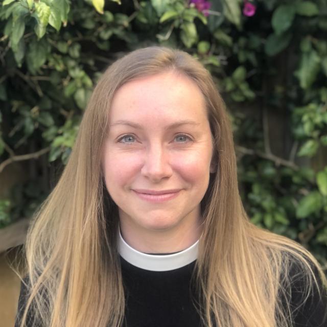 Grace Flint smiles directly at the camera, standing in front of a leafy background. She is a white woman with dirty blonde hair, blue eyes, and a pinkish complexion. She wears a neckband collar in deference to her role as an Episcopal priest.