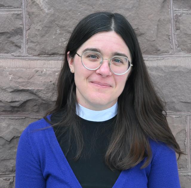 Jessica Frederick softly smiles at the camera with her head tilted to the left as she stands in front of a stone wall. She is a white woman with dark hair parted in the center and a pinkish complexion. She wears round glasses, stud earrings, and a cobalt blue cardigan over her neckband top, in deference to her status as a transitional deacon in the Episcopal Church.