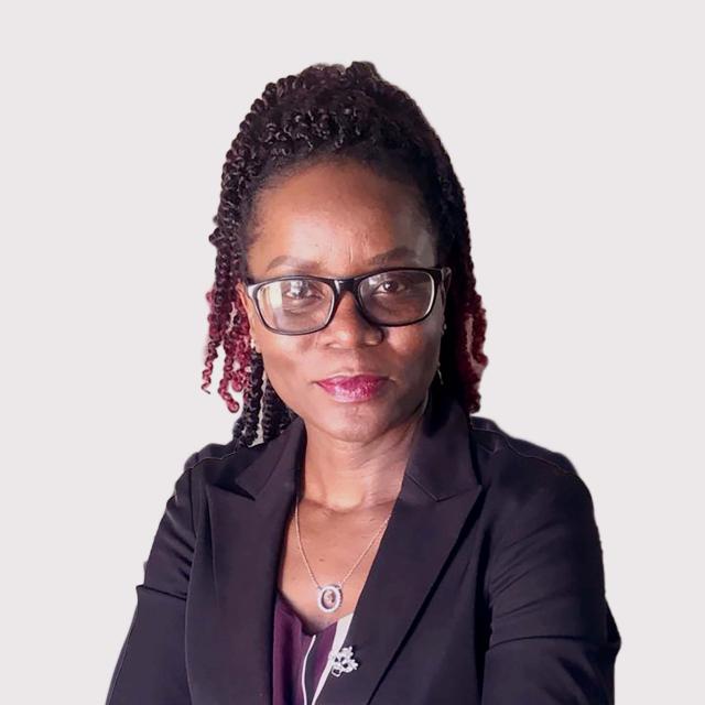 Kudzai Belinda Deve softly smiles at the camera in front of a white background. She is a Black woman with a burgundy ombre twists, wearing a black blazer, eggplant blouse, rectangular-framed glasses, and silver pendant necklace.